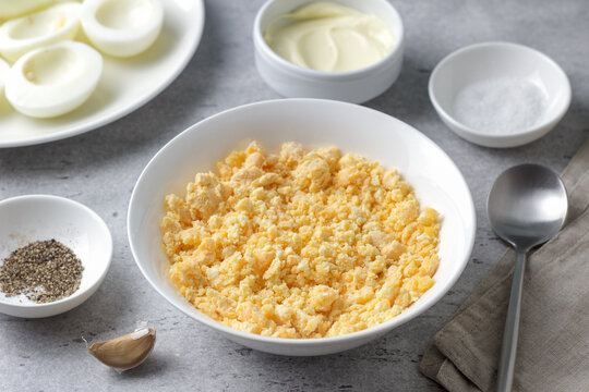 Crumbled hard boiled egg yolks in a bowl, garlic, mayonnaise and seasonings, prepared as filling for stuffed eggs on a gray textured surface