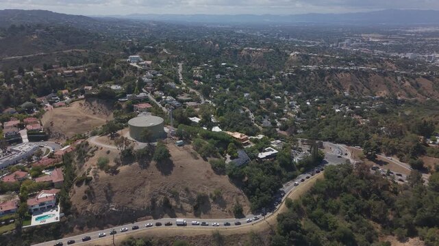 Dry chaparral hillsides with water tank and winding canyon road with traffic overlook sprawling San Fernando Valley under hazy skies in Sherman Oaks, California, USA. Aerial drone flyover shot.