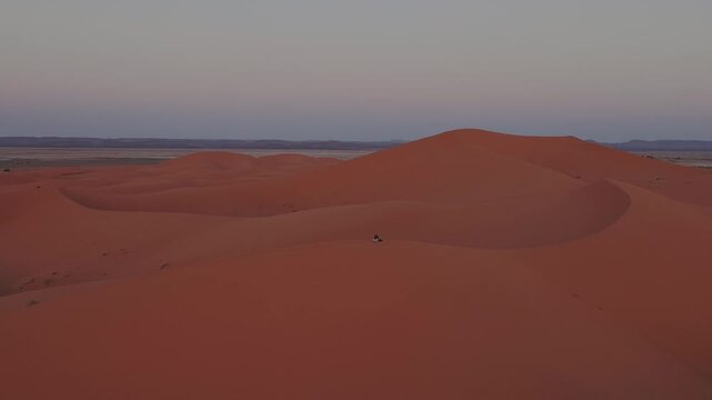 Lone traveler stands on a dune crest in Erg Chebbi surrounded by immense Sahara formations, emphasizing scale, solitude and vast desert wilderness, drone parallax shot.