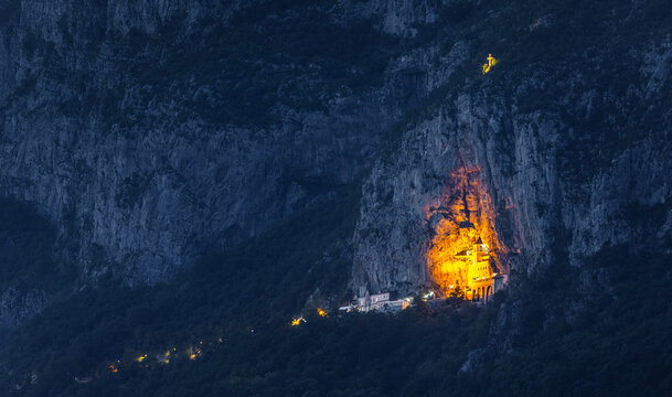 View of Manastir Gornji Ostrog illuminated against the dark cliffside at dusk, featuring warm golden lights glowing from the monastery architecture in Dabovici, Opstina Danilovgrad, Montenegro.