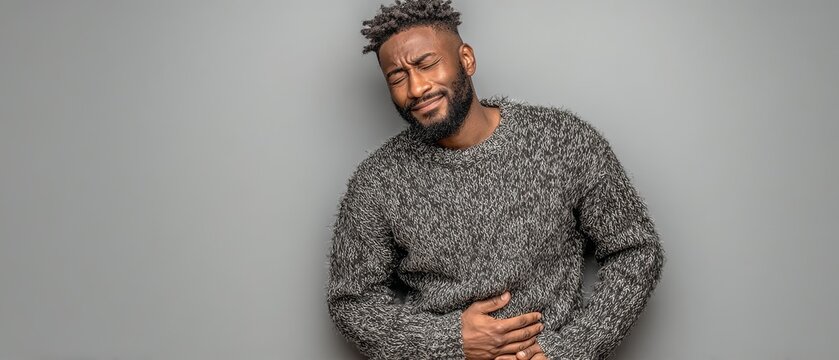 A man in a gray sweater grimaces in pain while clutching his stomach against a neutral background, illustrating acute physical discomfort.