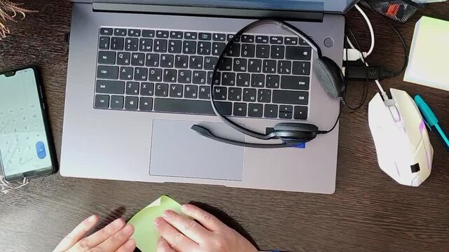 Overhead view of a woman writing notes at a desk with laptop, headset and smartphone while working from home. Remote work, home office, technology, productivity, business.