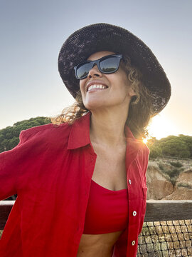 Smiling woman in a black sun hat and sunglasses wearing a red shirt and crop top at sunset on a coastal boardwalk, enjoying warm light, summer vacation, confidence and relaxed happiness.