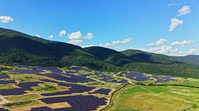 A wide view shows solar panels laid out across fields in a valley. Mountains are visible in the background under a bright blue sky with some clouds.