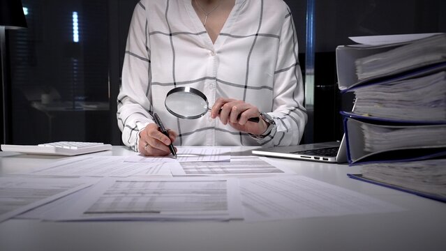 Professional woman uses magnifying glass and pen to scrutinize financial reports and documents on an office desk, symbolizing auditing, investigation and detailed data analysis