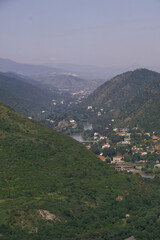 Fototapeta premium Scenic valley town nestled between lush green mountains under a hazy sky.A picturesque view of a small town situated in a verdant valley, surrounded by rolling hills and mountains, with a river flowi