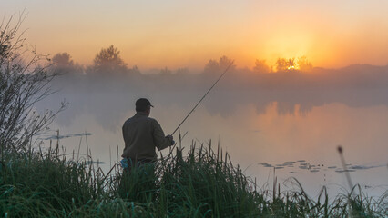 fisherman stands by a misty lake at sunrise, holding a fishing rod against a golden, hazy sky background © Александр Арендарь