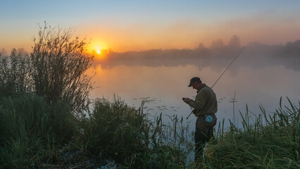 A lonely fisherman stands by a misty lake at sunrise, holding his rod while golden light reflects on the calm water © Александр Арендарь