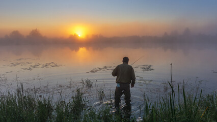 A lonely fisherman stands in the calm water during a golden sunrise, surrounded by thick mist over a peaceful lake © Александр Арендарь