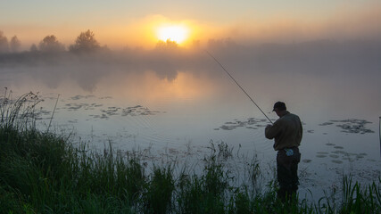 fisherman stands by a misty lake at sunrise, holding a fishing rod while golden light reflects on water © Александр Арендарь