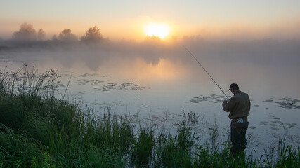 fisherman stands by a misty lake at sunrise, holding his rod while golden light reflects on the calm water © Александр Арендарь