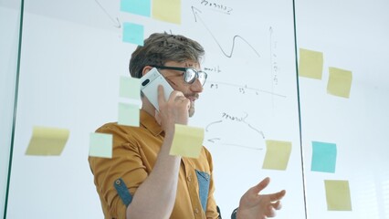 Smiling businessman on a phone call mapping ideas on a glass wall with colorful sticky notes, organizing tasks and brainstorming strategy in a modern office for project planning © Hanna