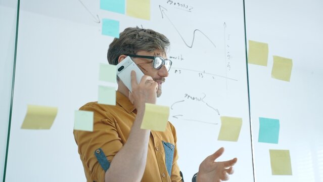 Smiling businessman on a phone call mapping ideas on a glass wall with colorful sticky notes, organizing tasks and brainstorming strategy in a modern office for project planning