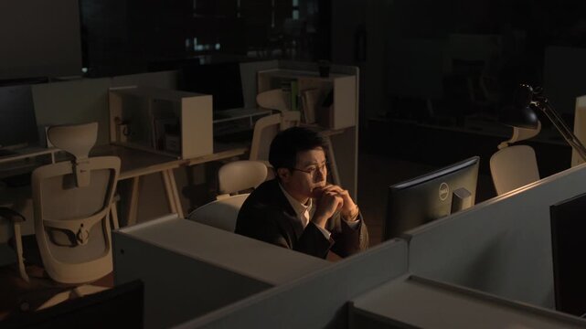 A businessman resting his chin on his hand in a thoughtful pose while sitting alone in a dark office late at night.