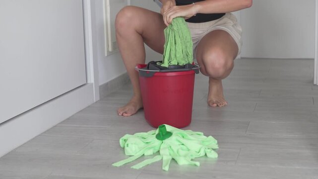 Woman replacing an old mop head with a new one during home cleaning. Close-up domestic scene with cleaning supplies, red bucket and microfiber mop on the floor in a modern apartment