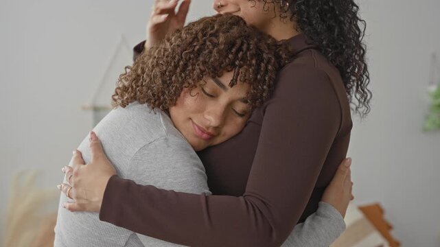 Woman embraces woman, hand cradling head and arms wrapped in hug on bedroom bed with pillows and wall decor; comfort support.