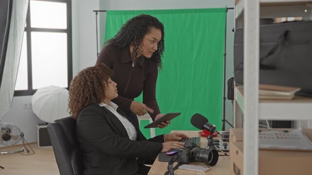 Woman standing and pointing at tablet while seated woman types on keyboard in studio with green screen and camera and microphone; collaboration mentorship.
