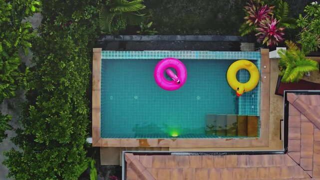 Private tropical villa pool in Phuket with bright flamingo and duck inflatable rings, blue water framed by lush green foliage. Summer vacation relaxing travel escape. Aerial top-down view drone shot
