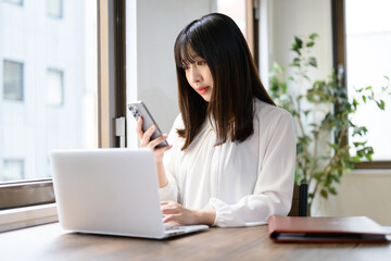 オフィスでノートパソコンとスマホを使って仕事をする日本人女性 © Yamatatsu
