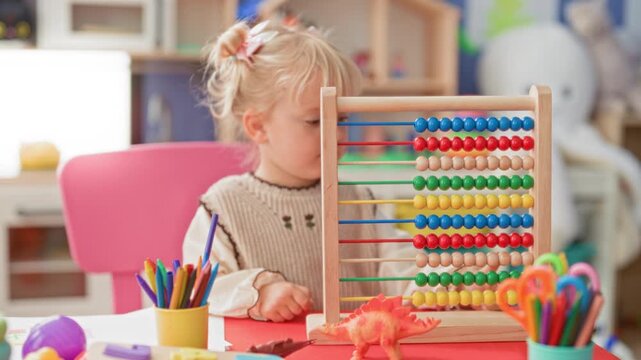 Little blonde girl counts and slides colorful beads on a wooden abacus with hand visible in a daycare building filled with crayons and toys; concentration learning play.