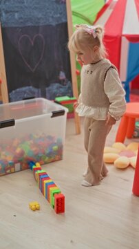 Little blonde girl jumps with hands raised over a row of colorful toy blocks near a clear storage bin and chalkboard easel inside a children's playroom building; childhood joy.