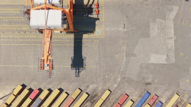 Aerial top-down view of a massive gantry crane lifting and moving a white cargo container across a port terminal yard, showcasing modern logistics and international trade operations