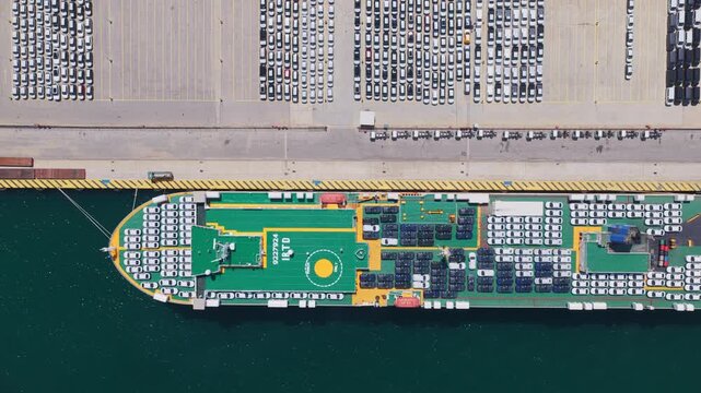 Top-down aerial view showing a massive roll-on roll-off vehicle carrier ship maneuvering to dock at a commercial port, with thousands of new cars parked on the pier ready for export