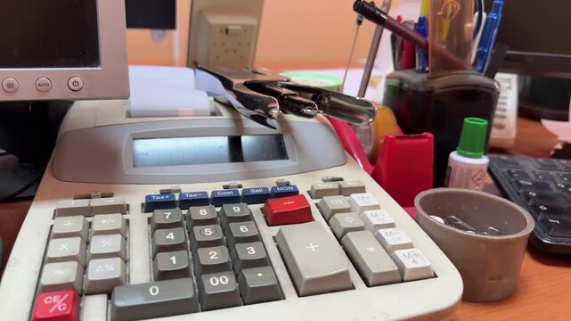 A printing calculator and binder clip sit on a cluttered office desk, with paper roll, pens, keyboard, monitor, correction fluid and paper clips visible.