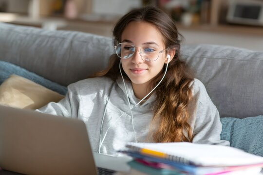 Teenage girl with glasses and earphones studying on laptop at home. Student watching online lesson. Distance learning concept