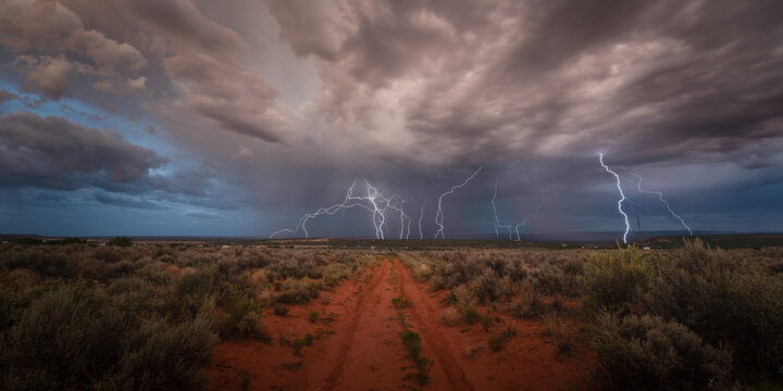 View of multiple lightning bolts striking the horizon over a red dirt road and desert shrubs under a dark stormy sky in Kanab, Utah, United States.