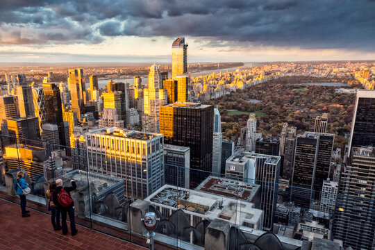 Tourists photographing the Manhattan skyline and Central Park at sunset from the observation deck, symbolizing urban travel and exploration, Top of the Rock, Rockefeller Center, New York City.