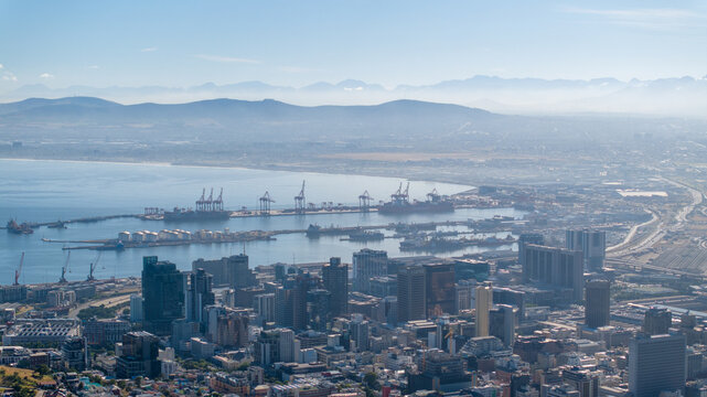 Aerial view of the Cape Town harbor with container ships, industrial cranes, and the dense urban skyline set against distant mountains in Cape Town, Western Cape, South Africa.