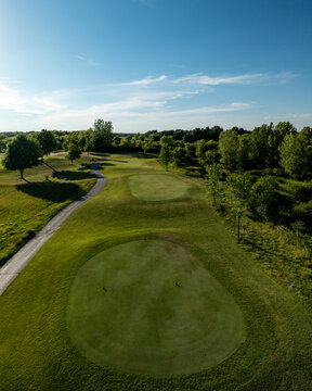 Aerial view of a golf course green with a winding path, lush green trees, and manicured grass under a clear blue sky in Lake Geneva, Wisconsin, United States.