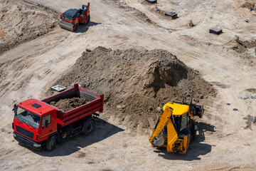 an active construction site shows a red dump truck and a yellow backhoe loader working near a large dirt pile, with a road roller compacting the sandy terrain in the background. © Andrey