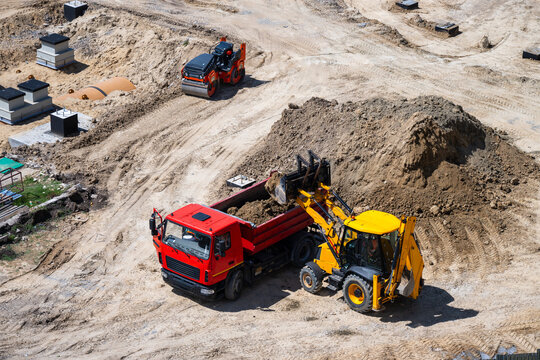 an active construction site captures a yellow backhoe loader in the act of dumping a scoop of earth into the bed of a waiting red dump truck, while a road roller sits on a dirt path in the background.