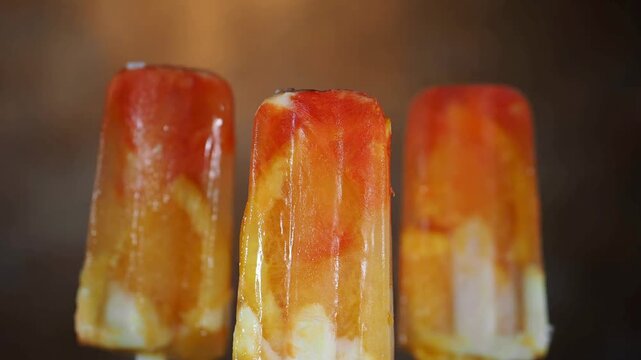 4K Video of Delicious Homemade Fruit Popsicles with Fresh Orange and Strawberry Pieces Melting Slowly in a Row Against a Dark Studio Background, Perfect Commercial Visual for Summer Refreshment