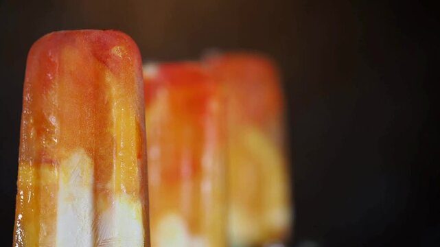 4K Video of Delicious Homemade Fruit Popsicles with Fresh Orange and Strawberry Pieces Melting Slowly in a Row Against a Dark Studio Background, Perfect Commercial Visual for Summer Refreshment
