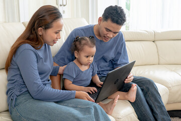 Fototapeta premium Asian Parents with Cute Toddler Daughter Watching Laptop Together on Sofa