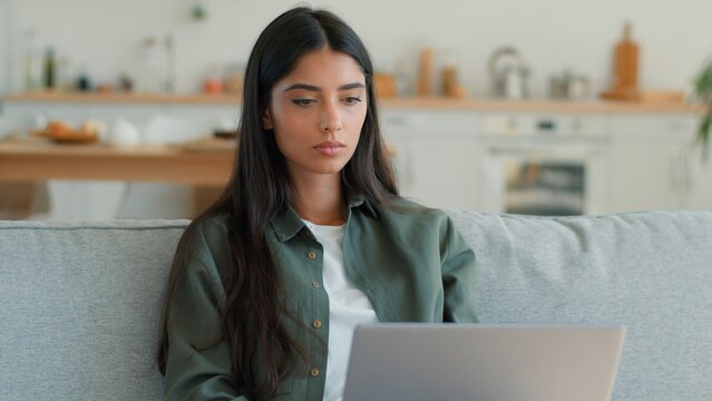 Concentrated Arabian Indian woman working on laptop at home couch focused freelancer student female Hispanic lady typing browsing internet remote work studying online shopping using computer PC indoor