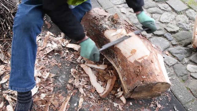 Skillful woodworker wearing protective gloves using traditional drawknife to carefully peel the rough bark from a large tree log, preparing the timber for a carpentry project