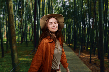 Fototapeta premium Woman in bamboo forest wearing straw hat and orange jacket walking on wooden boardwalk path, smiling at camera. Nature portrait, travel, leisure, peaceful getaway and outdoor lifestyle.
