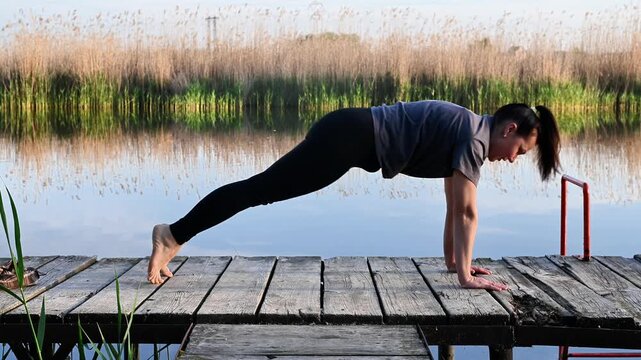 Woman performs plank pose on wooden pier. pushup exercise on old bridge near river shore. lady enjoys fitness and stretching activity for mental wellness.female person does yoga stretch during sunrise