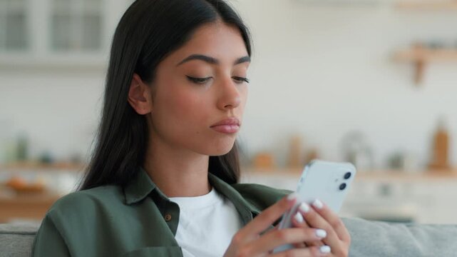 Shocked surprised Indian Muslim woman using mobile phone at home sofa looking at cellphone confused worried news scrolling social media app Arabian female girl puzzled thinking browsing device indoors