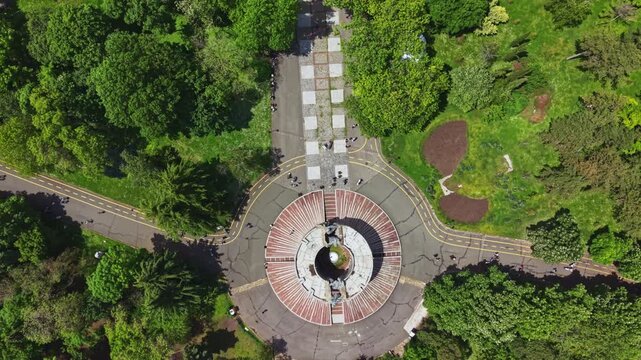 Overhead fountain winding path through trees, drone glide revealing radial plaza opening into shaded walkway, textured paving, dense shrubs, small groups