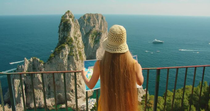 Back view of tourist woman with map facing Faraglioni cliffs from Capri lookout terrace