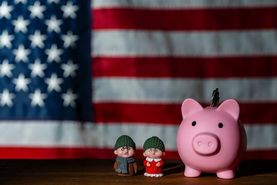 Pink piggy bank with elderly couple miniatures on wooden table against a blurred USA flag. Represents American retirement savings, social security, senior finance, and pension funds