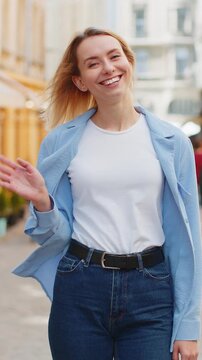 Cheerful young woman tourist smiling friendly at camera, waving hands gesturing invitation hello, hi, greeting, goodbye, welcoming with hospitable expression outdoors. Lady on urban city town street