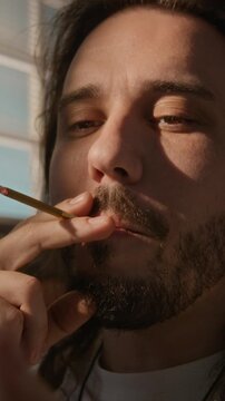 Vertical video of young adult Caucasian man smoking hand-rolled cigarette and exhaling thick cloud of smoke, relaxing at home. For campaigns about substance abuse, addiction, or mental health
