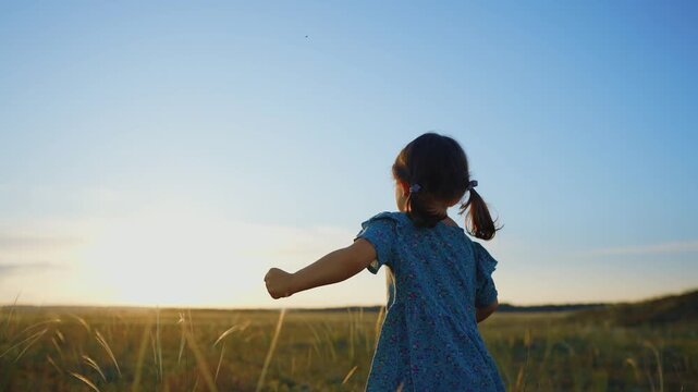 A girl is running through a field of grass during sunset. The girl appears joyful and carefree, moving towards the horizon under a clear sky.
