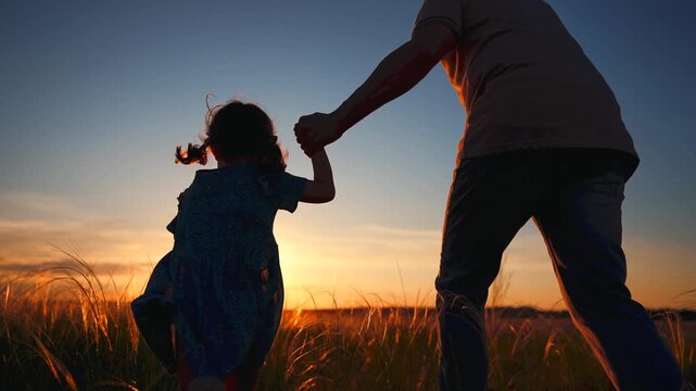 A man and a girl are running hand in hand through a field as the sun sets. The man and girl enjoy the warm light of the sunset while the grass sways around them.